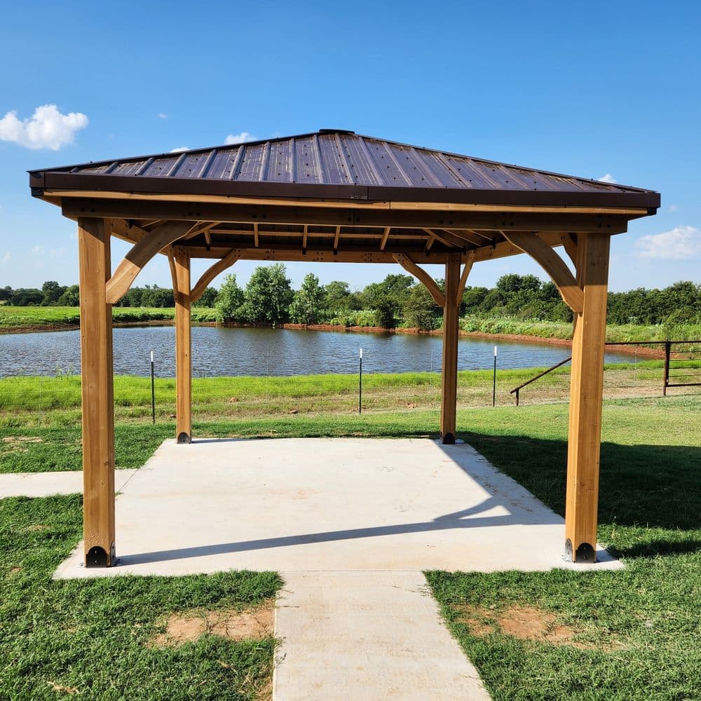 Wooden gazebo with a metal roof by a serene pond, surrounded by green grass and trees.