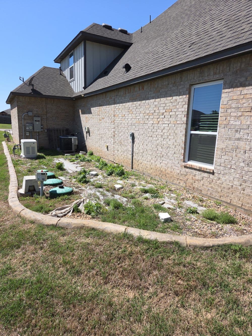 D yard with overgrown plants, stones, and air conditioning units beside a brick home.