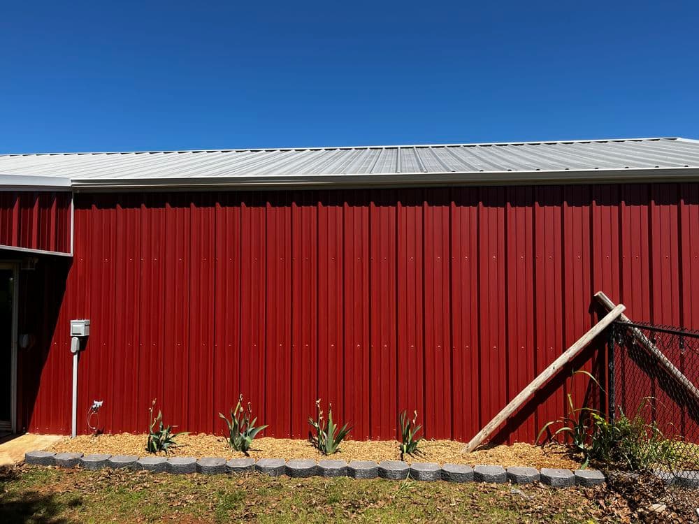 Red metal barn with grey roof, landscaped border, and clear blue sky in the background.
