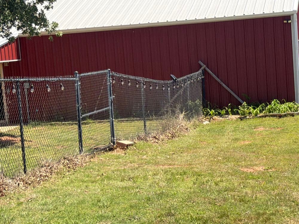 Chain link fence with rustic red barn and green garden in background on sunny day.