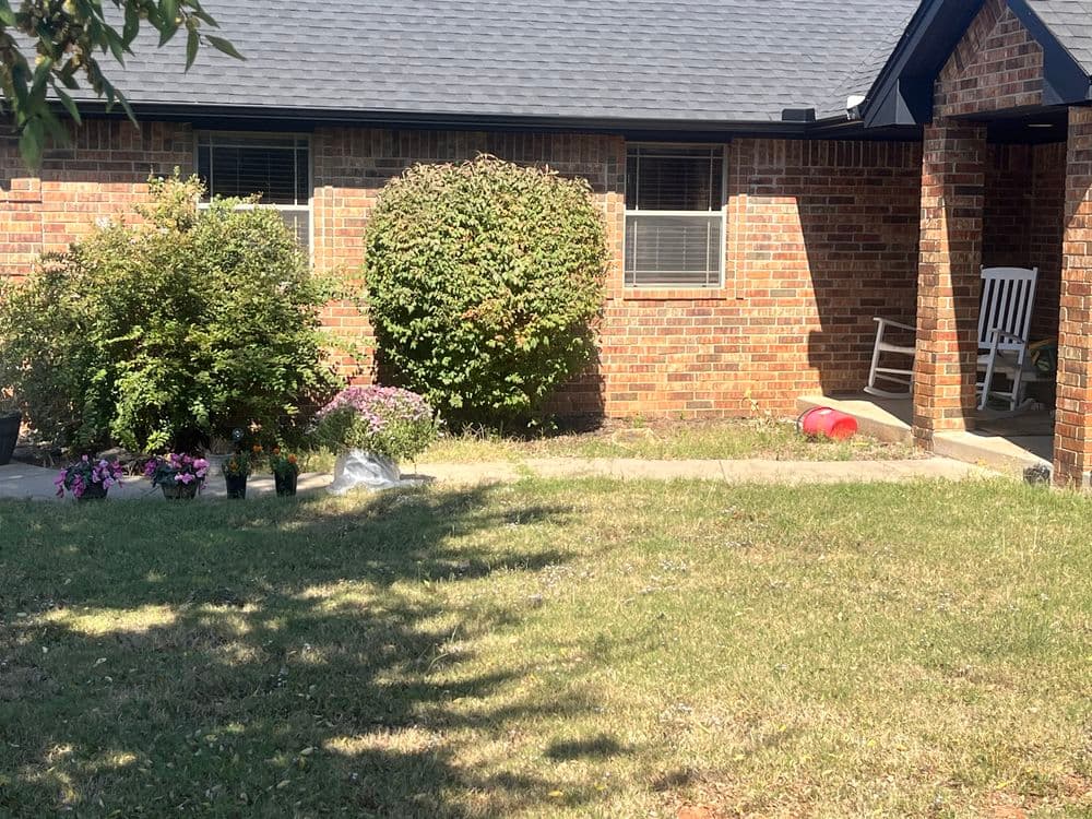 Brick house exterior with green bushes, flowers in pots, and a front porch chair.