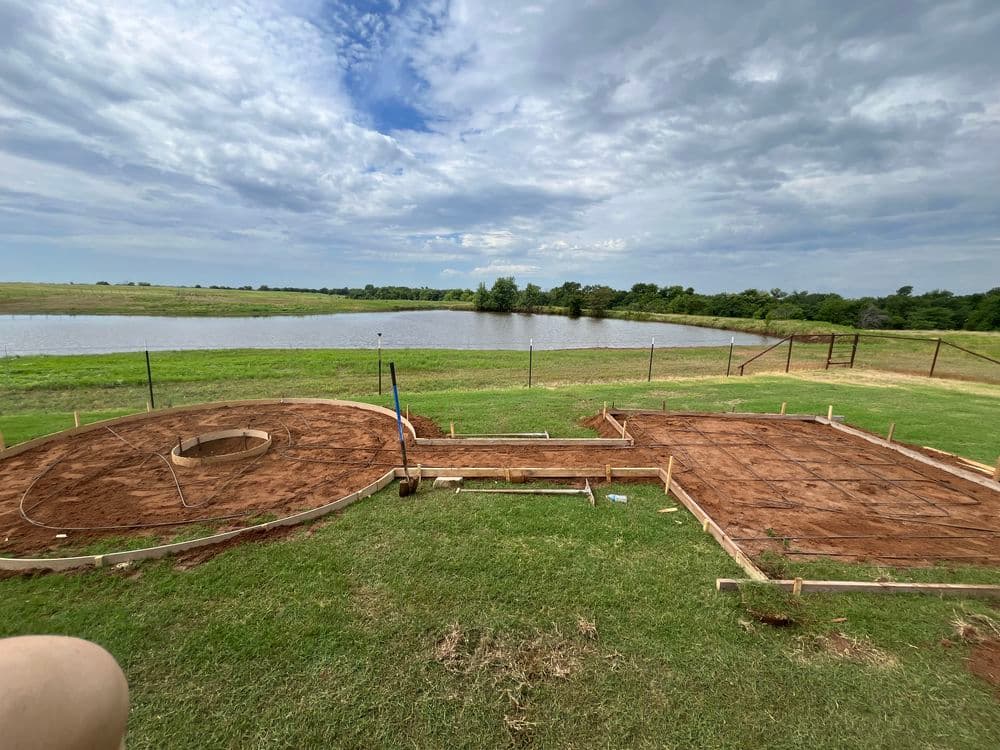 Construction site with a foundation laid on earth, overlooking a pond and green landscape.