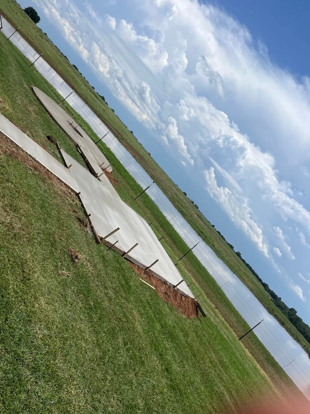 Damaged concrete slab near a body of water with green grass and blue sky.