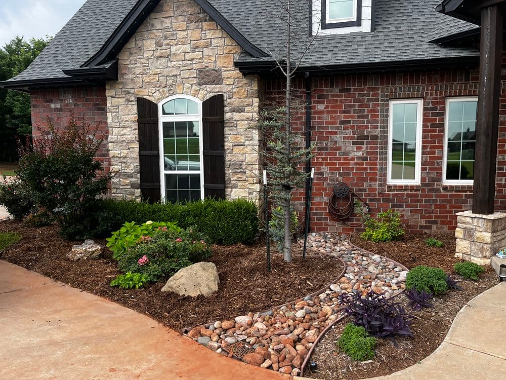 Landscaped home exterior featuring stone and brick facade, garden beds, and decorative rocks.