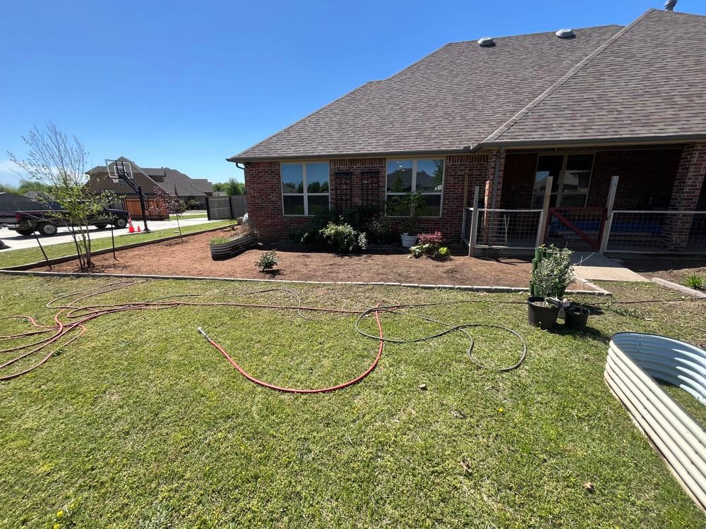 Landscaped backyard with new plants, mulch, and garden hoses next to a brick house.