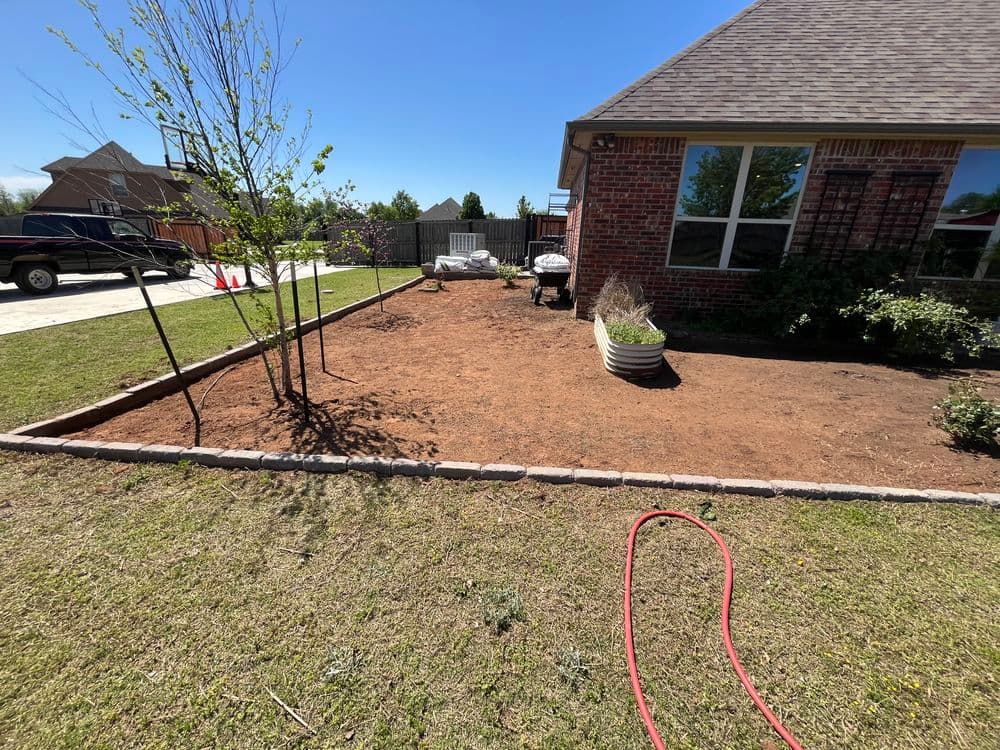 Landscaped yard with fresh soil, trees, and seating area beside a brick house on a sunny day.