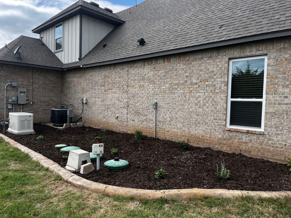 Backyard landscape with mulch, plants, and utility equipment near a brick house exterior.