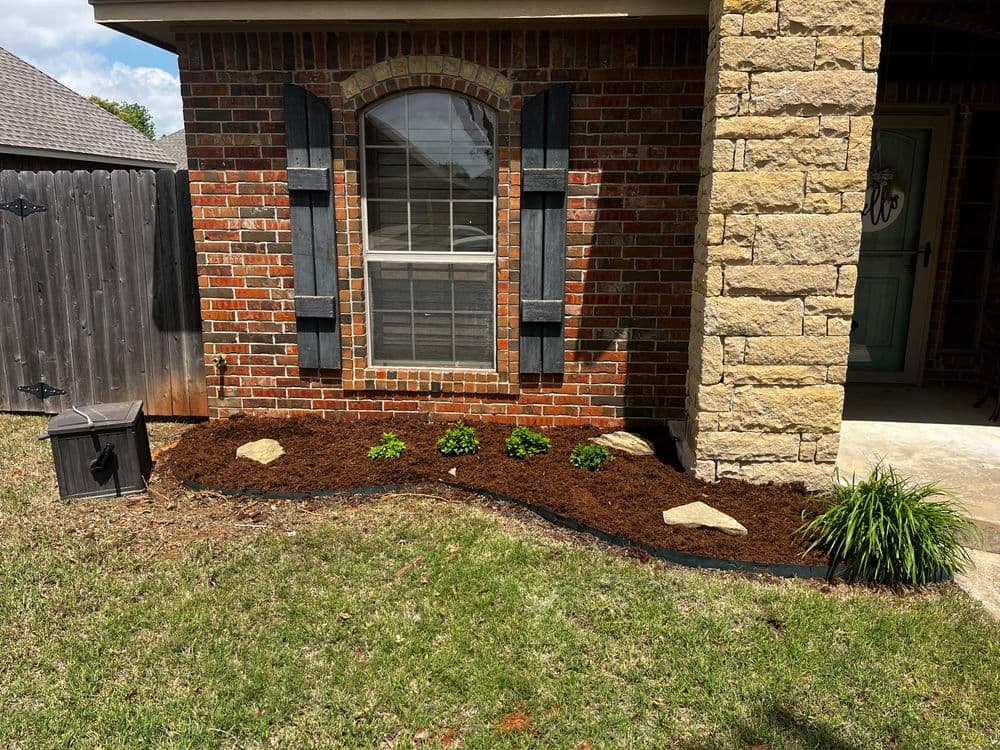 Freshly mulched garden bed with rocks and green plants near a brick house exterior.
