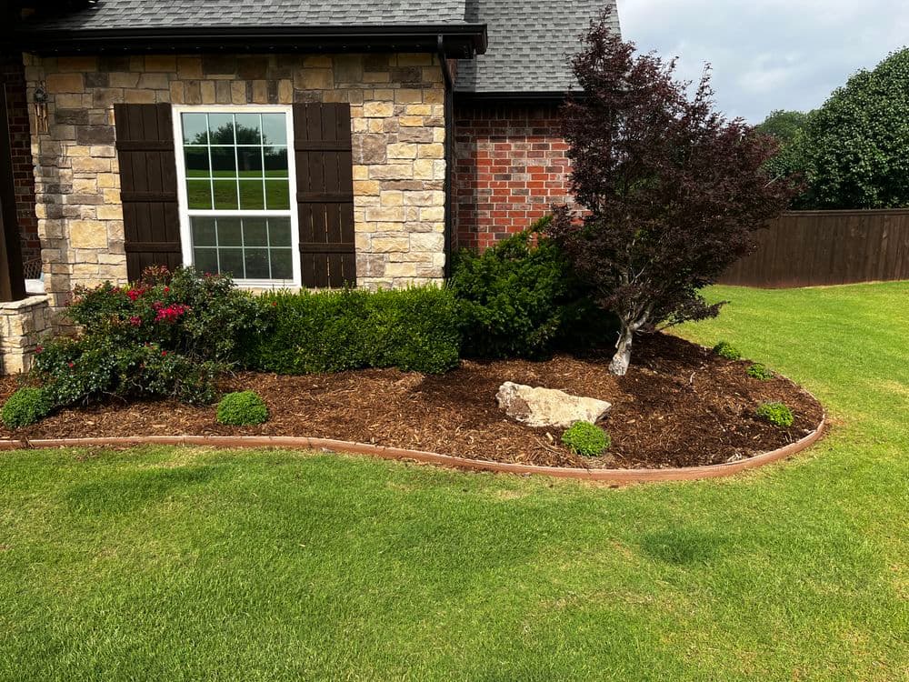 Landscaped garden with stone borders, colorful plants, and a small tree beside a brick house.