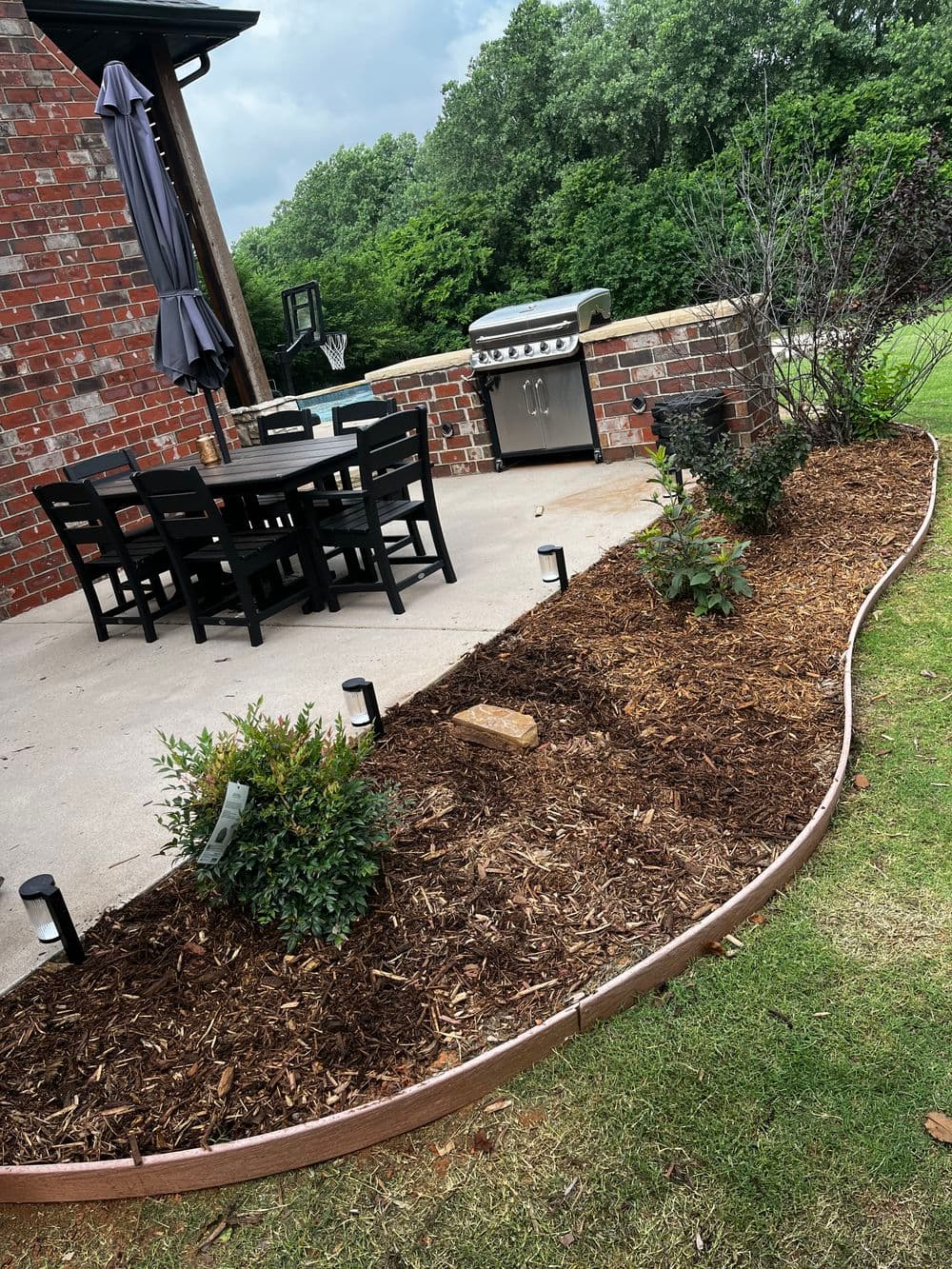 Backyard patio with a dining set, grill, and landscaped garden featuring mulch and small bushes.