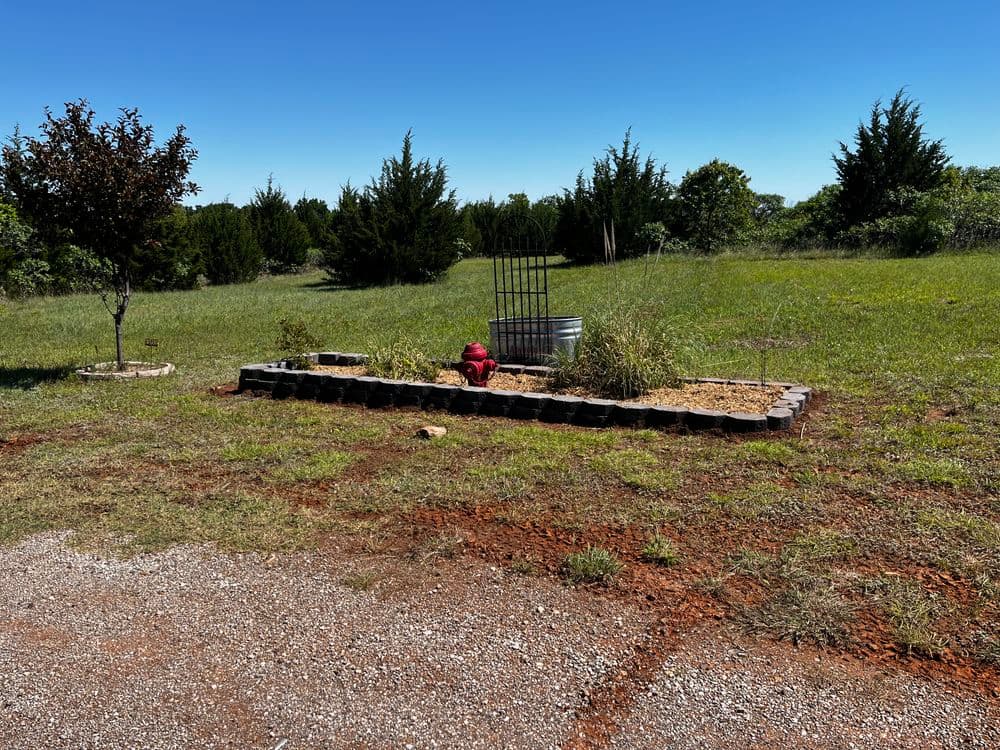 Landscape with stone flower bed, small tree, and gate in a grassy field under blue sky.