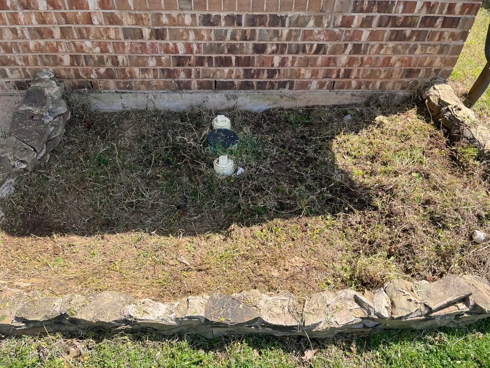 Overgrown garden bed with stone edging and two water valve covers against a brick wall.